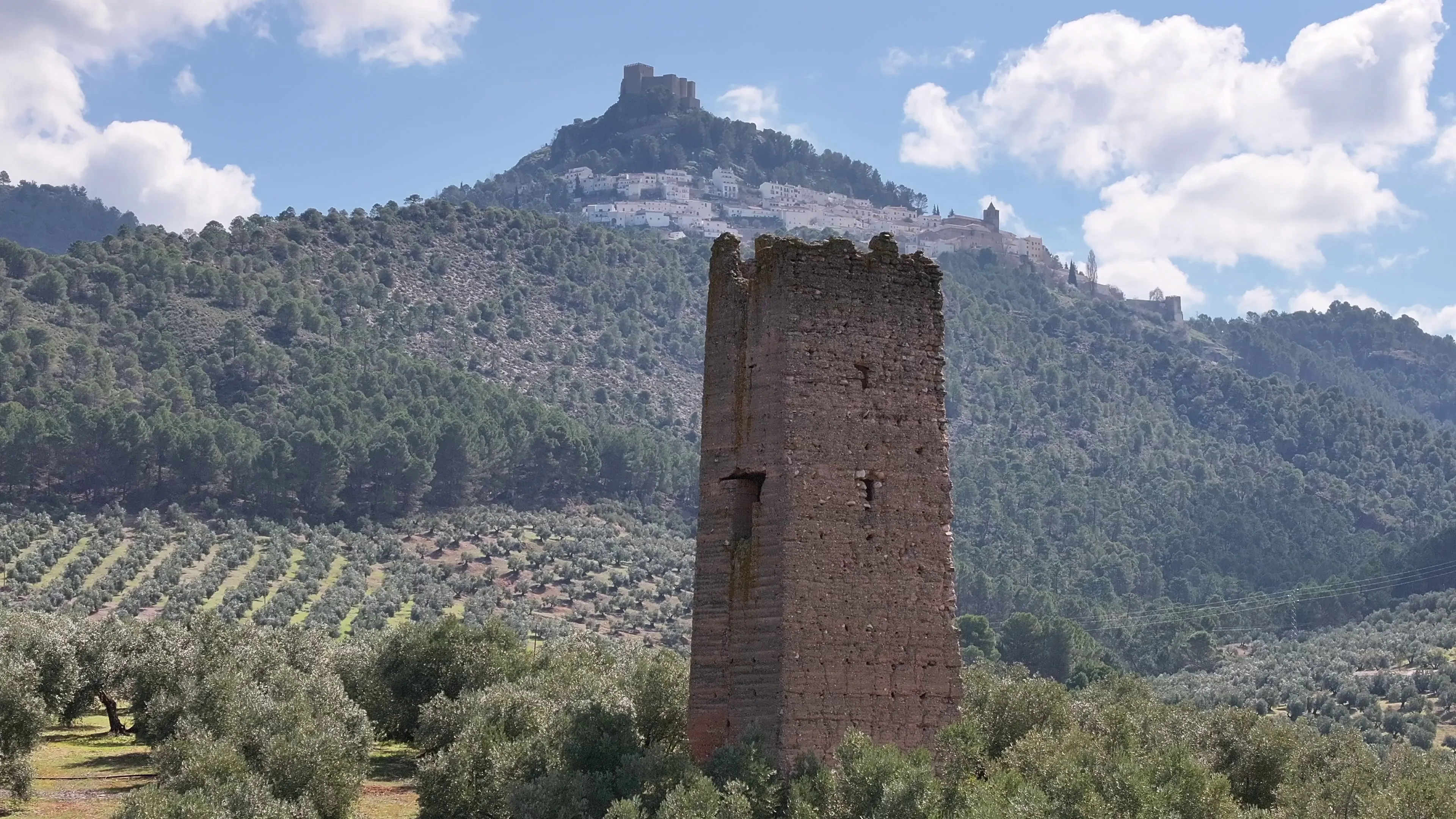 Torre entre olivares con un castillo en lo alto de la sierra al fondo